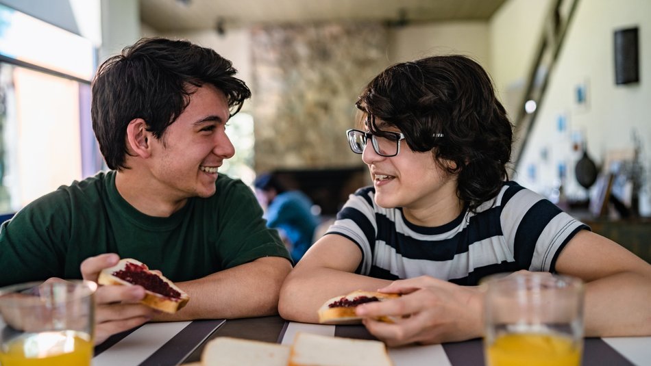 Two brothers smiling at each other as they sit at the table eating toast