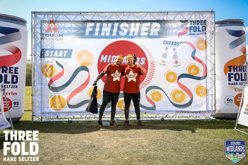 Two men at Tough Mudder in Action for Children t-shirts, pose in front of a large sign that says 'finisher'