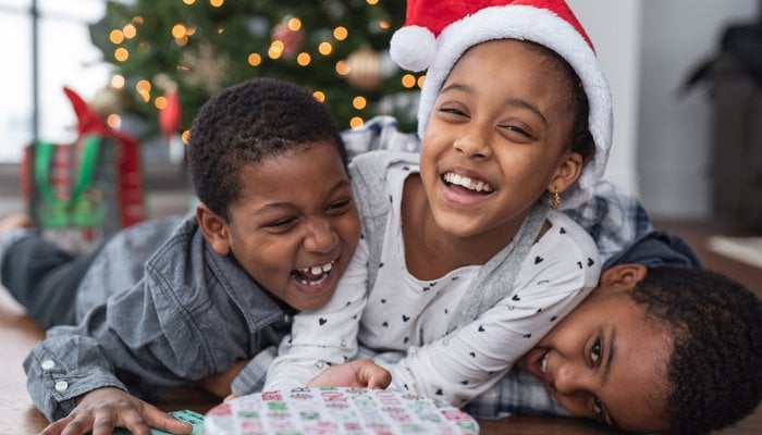 Three siblings laughing and laying on top of each other with Christmas gift presents in front of them a lit up tree in the background
