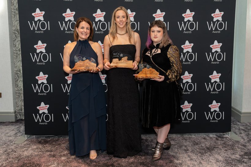 Three women in formal attire stand smiling with their wooden trophies in front of an Action for Children “WOI Awards” backdrop