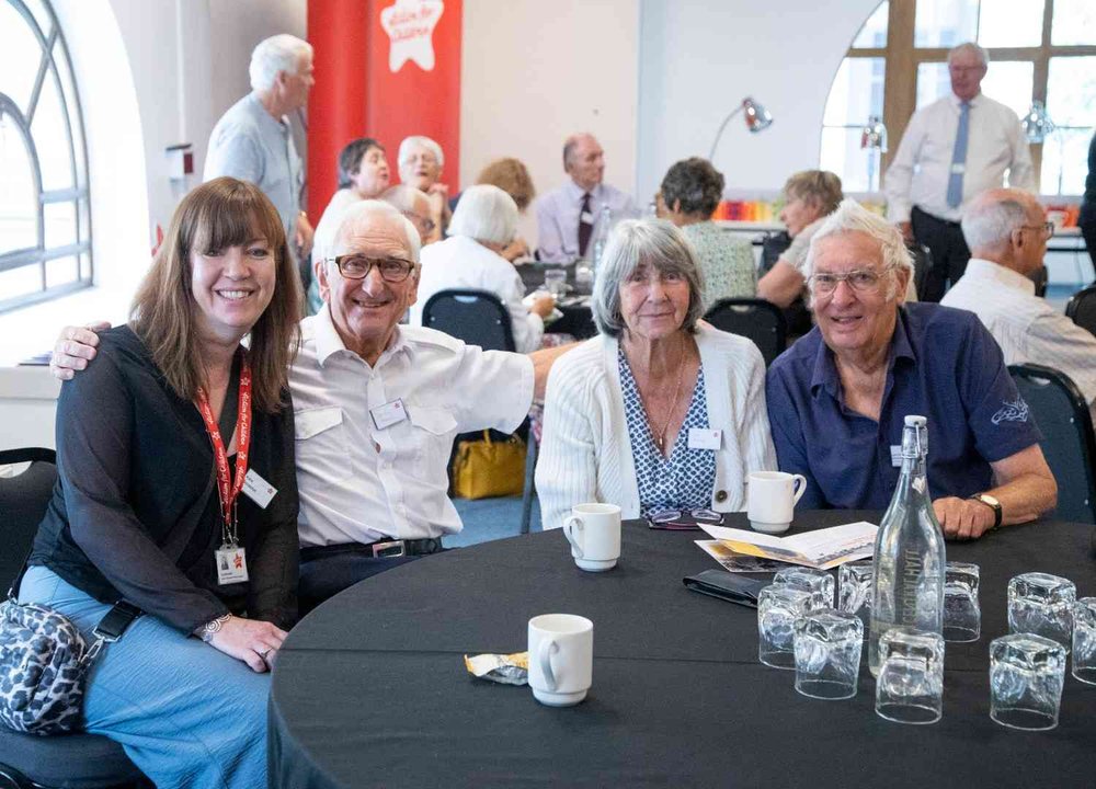 Group of older men and women sat at a table, arms around each other's shoulders, smiling