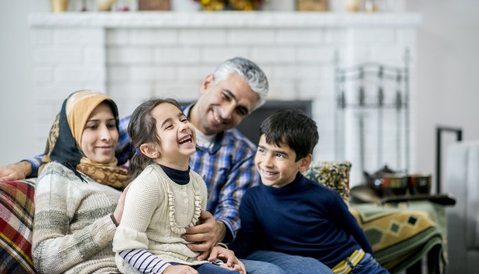 A Muslim mother, father, son and daughter are indoors in a living room. The parents are sitting on the sofa, and the daughter is laughing while being tickled.