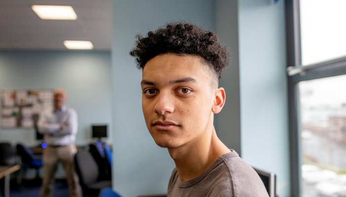A teenage boy sits in class and looks at the camera as his tutor stands in the background. He is wearing casual clothing.