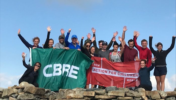 A group of adults with their hands in the air standing behind a stone wall holding a green CBRE flag and a red How Action for Children Works flag