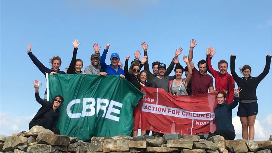 A group of adults with their hands in the air standing behind a stone wall holding a green CBRE flag and a red How Action for Children Works flag