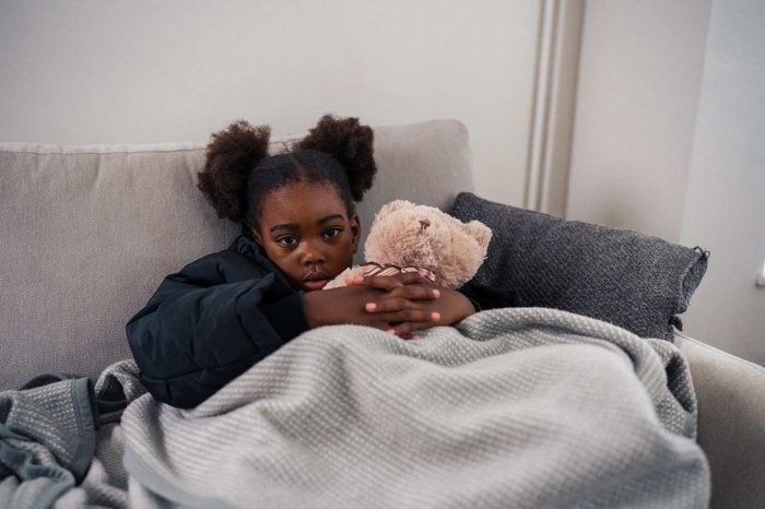 Girl huddled under blanket with teddy bear looking sad