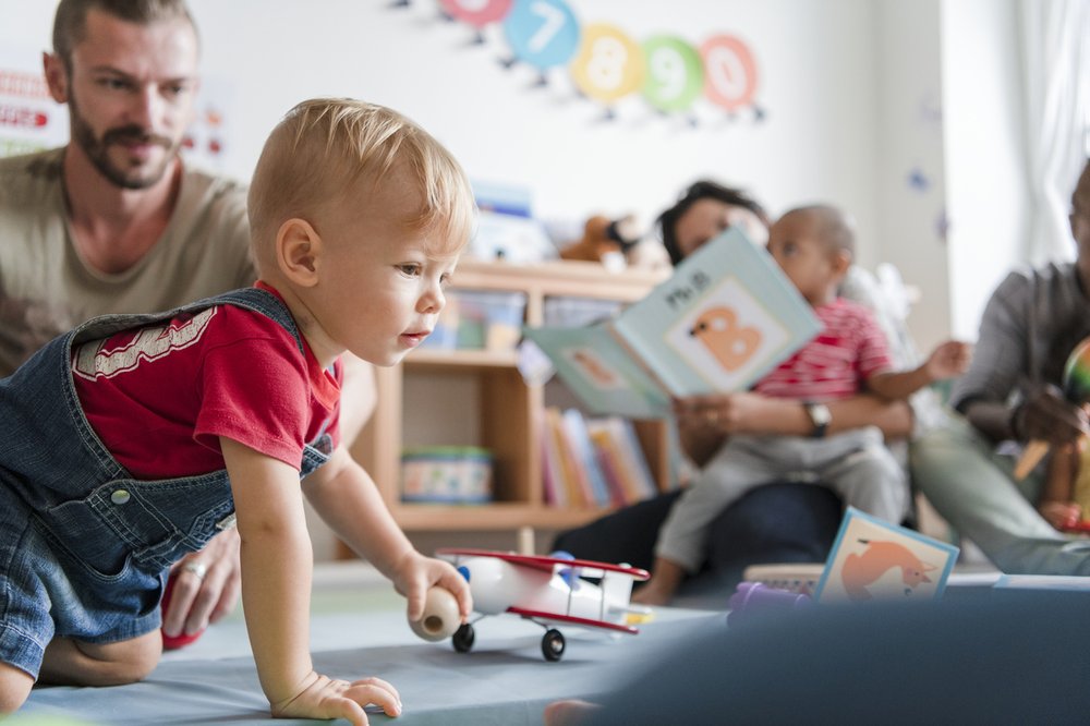 Baby boy crawling across a classroom floor while his father watches