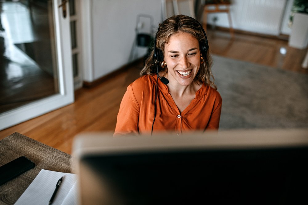 Cheerful young woman in headphones video conferencing on computer at home office