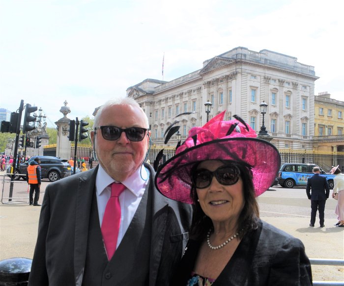 Portrait of foster carers Derrick and Sue are smartly dressed, smiling in front of Buckingham Palace