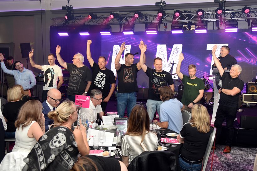 Eight men with their hands in the air standing in front of a stage at a music quiz