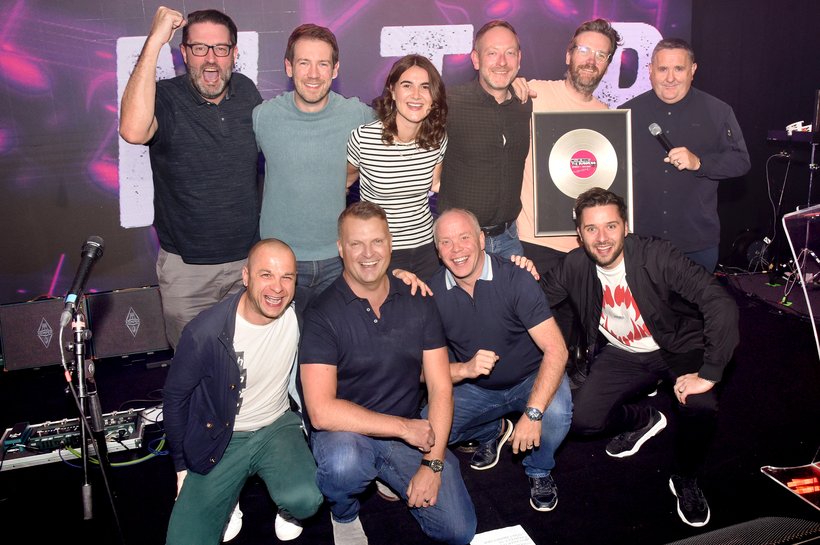 Nine men and one female on a music stage smiling at the camera, one holding a framed record
