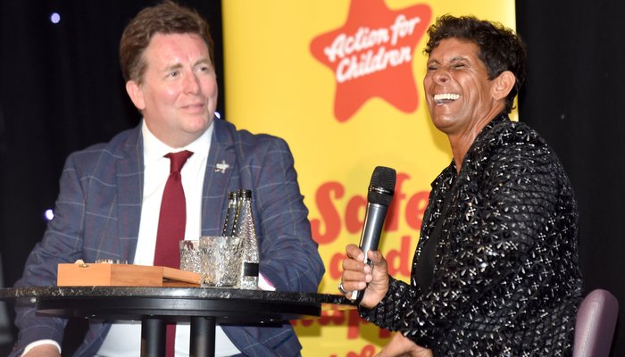 Fatima Whitbread sat on stage with a man in front of a yellow Action for Children pull up banner