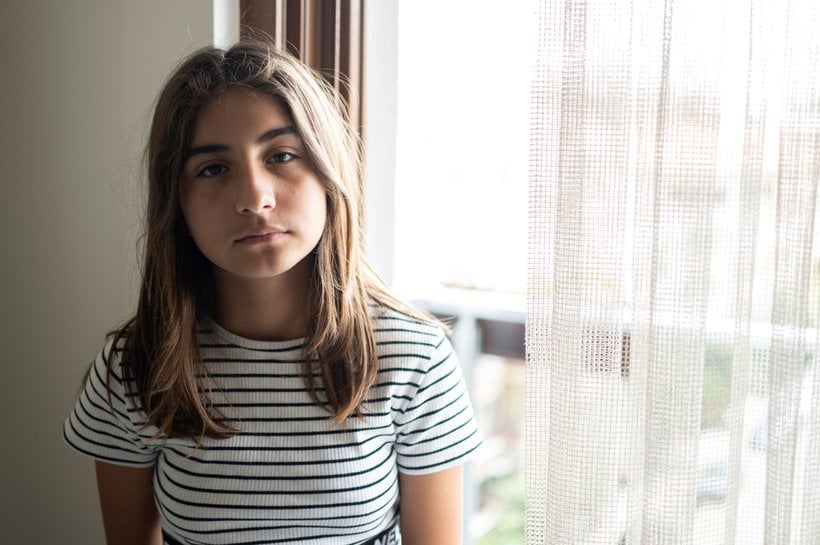 Girl standing up looking sad into camera next to curtains