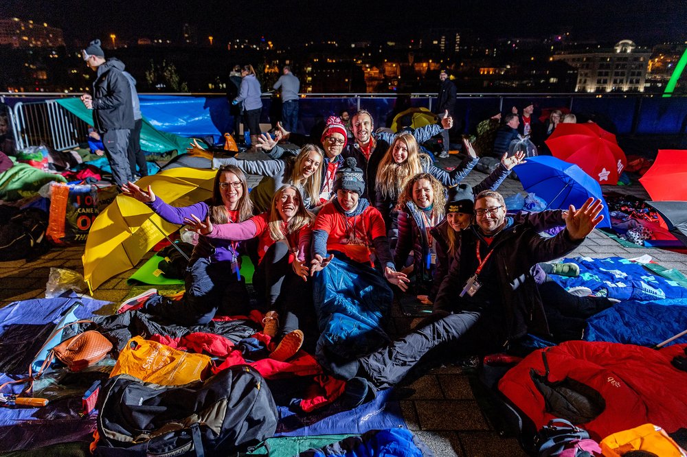 Group of happy, smiling people sat on the floor outside, surrounded by sleeping bag and umbrellas at a Byte Night sleep out event