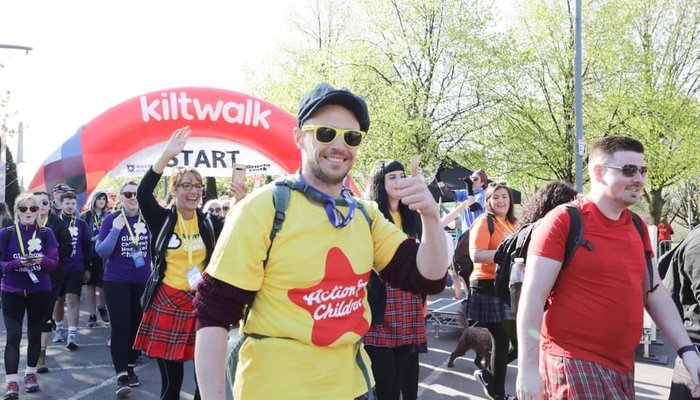 Group of Kiltwalkers walking, a man in an Action for Children t-shirt smiles and gives a thumbs up to the camera