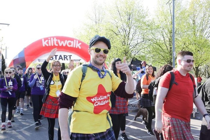 Group of Kiltwalkers walking, a man in an Action for Children t-shirt smiles and gives a thumbs up to the camera