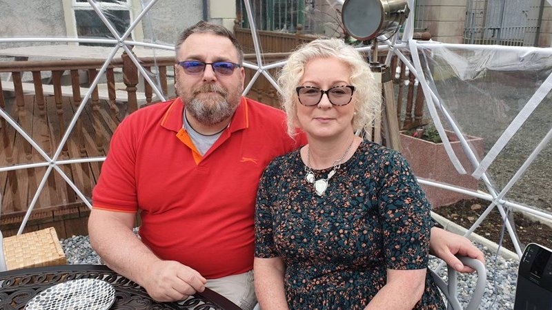 Foster carers John and Geraldine sitting close together at a table outside their home