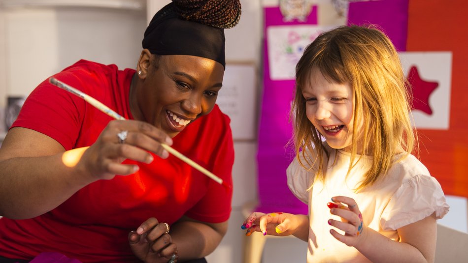 Abby, a young girl, is doing therapy play crafts with her support worker. They are both smiling and laughing.