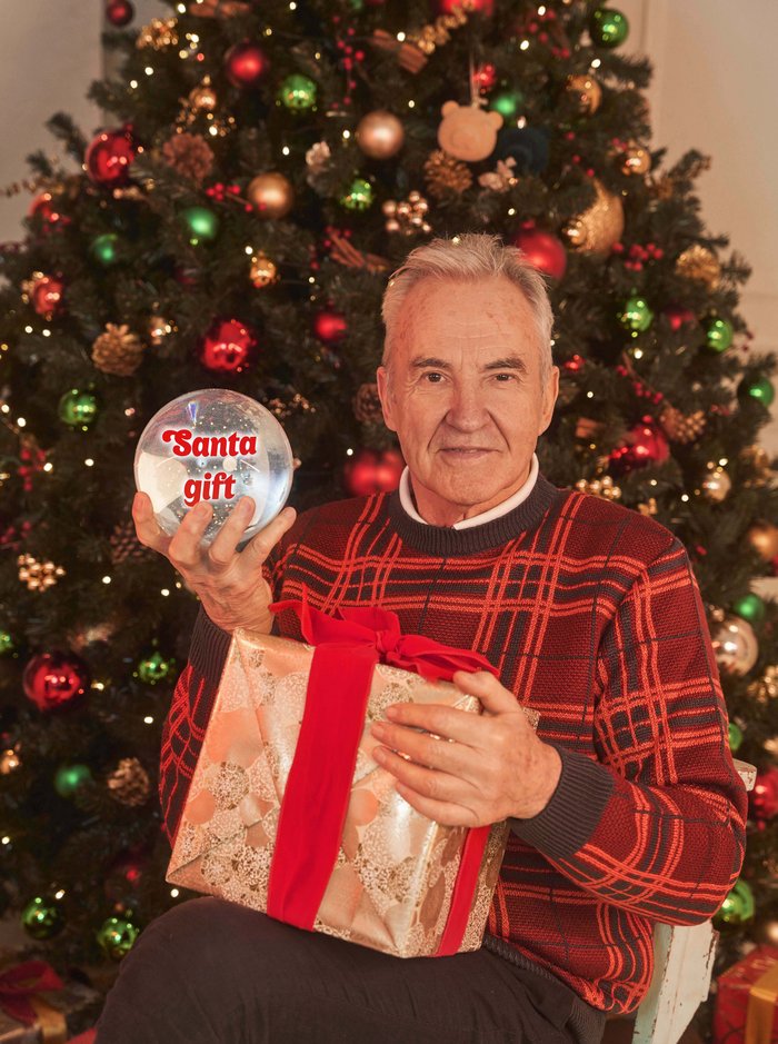 Larry Lamb sat in front of Chrsitmas tree holding a wrapped present and Snow globe that says 'Santa gift'