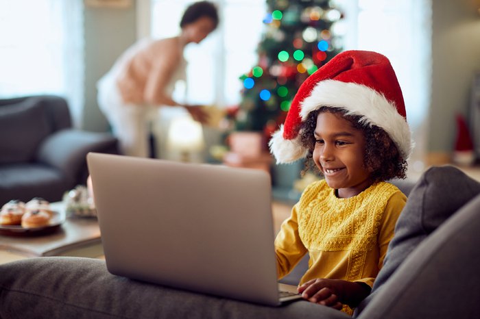 Little girl wearing Santa hat and using laptop while relaxing at home for Christmas. Her mother is in the background