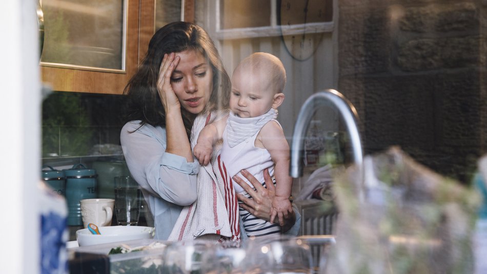 Stressed young mother in kitchen carrying baby on her hip