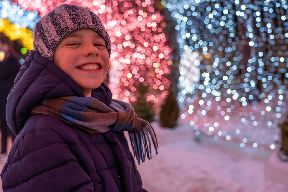Teen boy outisde in warm clothes smiles at the camera. He's infront of a a background of glowing Christmas decorations