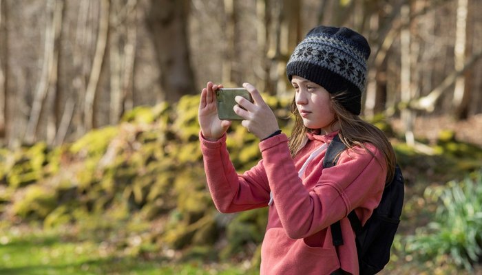 Teenage girl wearing a knit hat, standing outside surrounded by nature. She is taking a picture of the view with a mobile phone.
