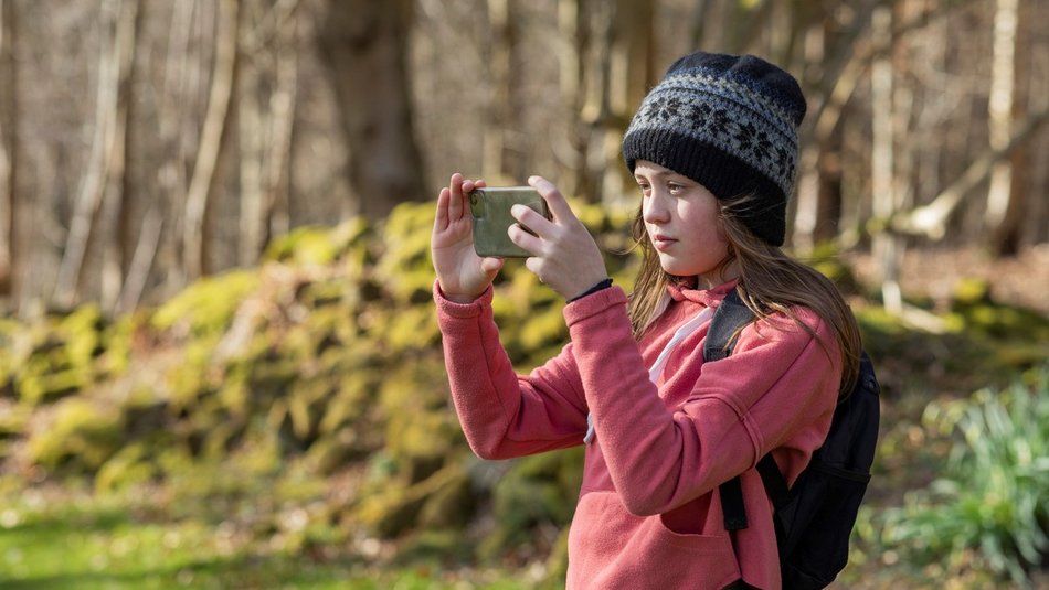 Teenage girl wearing a knit hat, standing outside surrounded by nature. She is taking a picture of the view with a mobile phone.