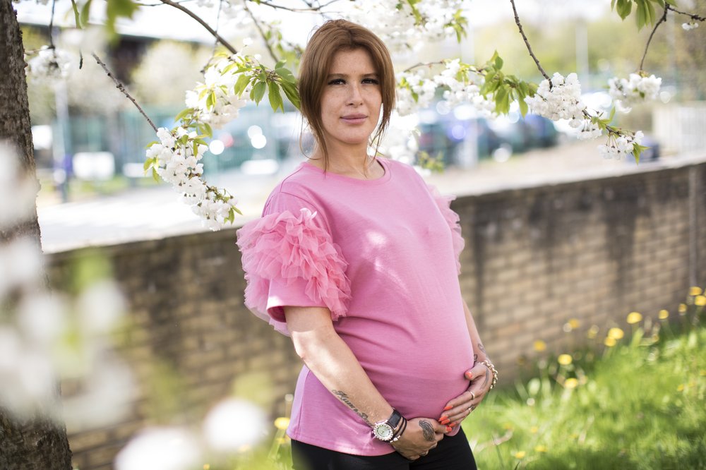 Victoria, a young woman in a pink top, poses holding her baby bump surrounded by flowering trees