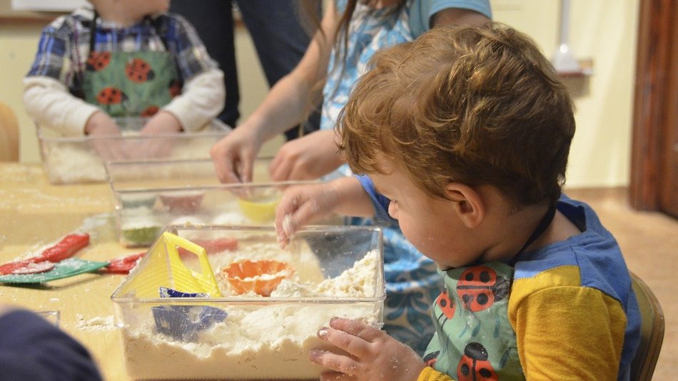Young boy sat at table playing with a plastic box filled with sand and small colourful toys. More children playing in the background