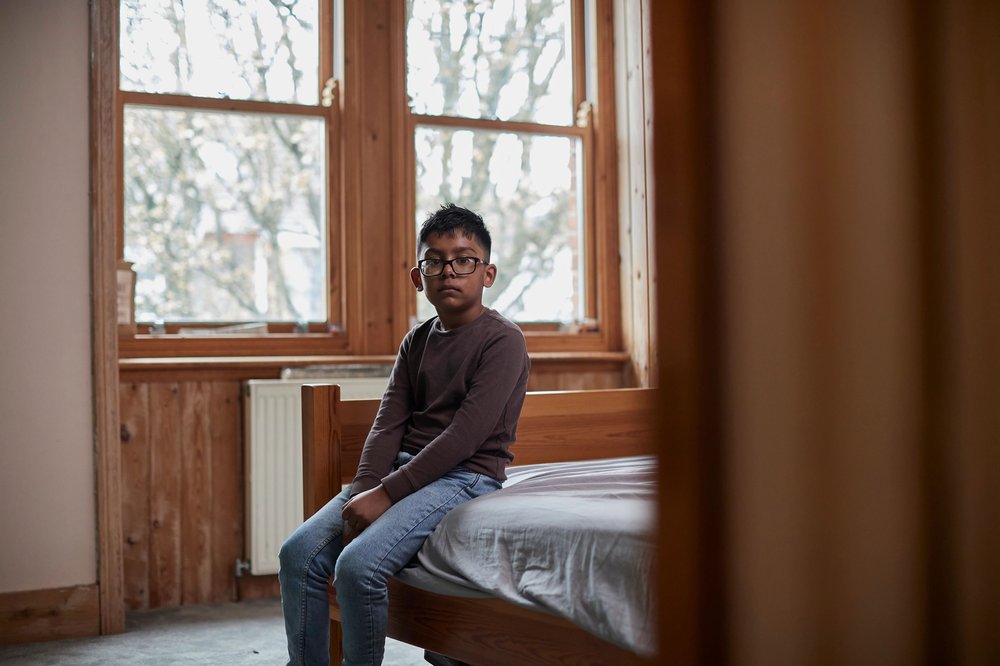 Young boy sat on edge of bed in an empty room looking at the camera