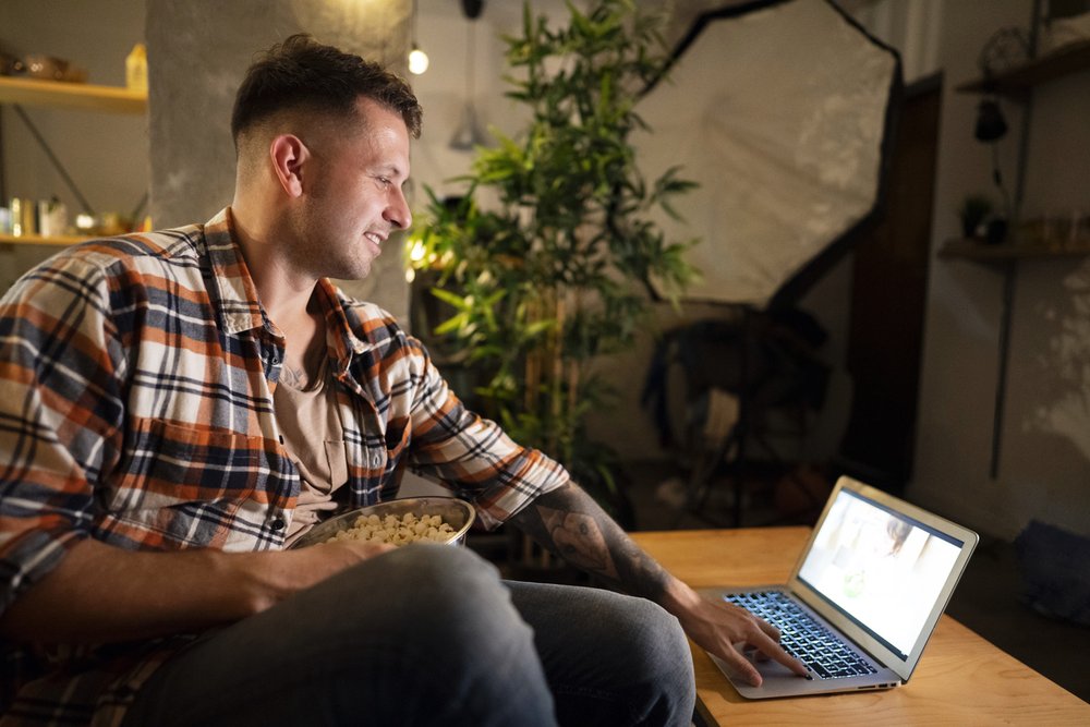 Young man playing a movie on his laptop and eating popcorn at  home