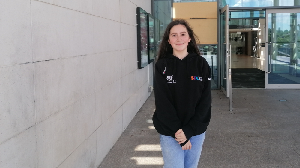 A teenage  girl, Kirsty,stands smiling in front of a building entrance. She wears a black hoodie and blue jeans